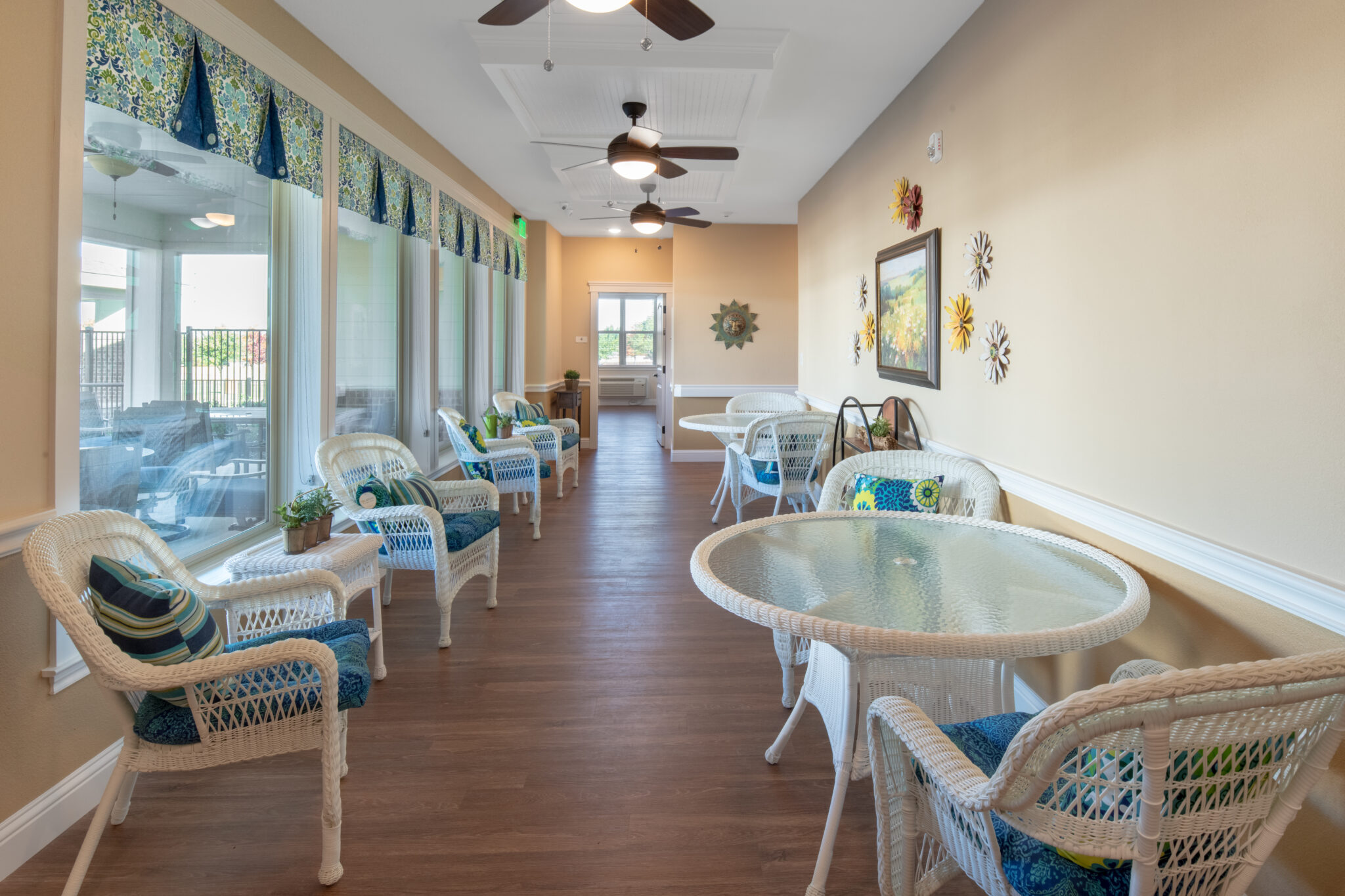 Sitting room in assisted living facility: Beige walls, wood floors, large windows for natural light, white wicker chairs, and wicker tables with glass tops.