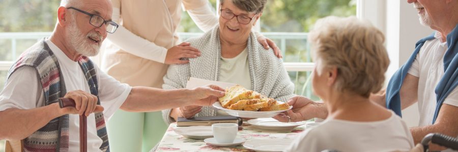 Group of senior friends with helpful carer sitting together at the table at nursing home dining room and eating cake
