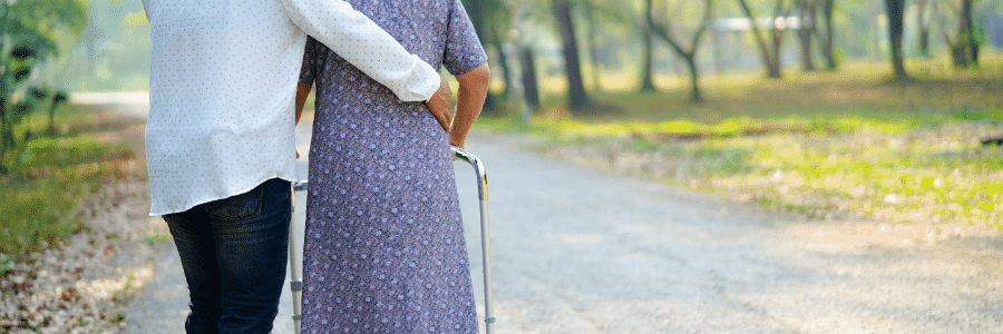 An elderly woman walks along a path with the support of a walker, accompanied by a younger woman who holds her waist, offering assistance.
